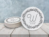 A stack of coasters are shown with a single coaster besides them on a white wooden surface. Coasters show Monogrammed Floral Wreath Initial design. This design has a floral wreath around a customizable large initial and date, all printed in black.