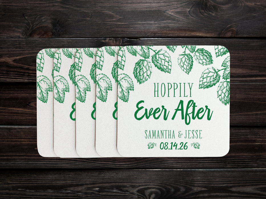 A stack of coasters are spread out across a dark wood surface. Coasters feature Hoppily Ever After design. This design uses green lettering and sketched drawings of beer hops. Wedding couples names and date can be customized.
