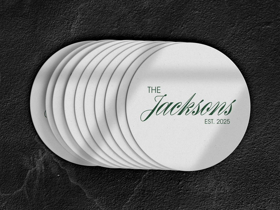 A stack of coasters are spread out on a dark black marble surface. Coasters feature Formal Last Name and Established Date design. This design has a white background and the happy couples last name and wedding year in emerald green writing.