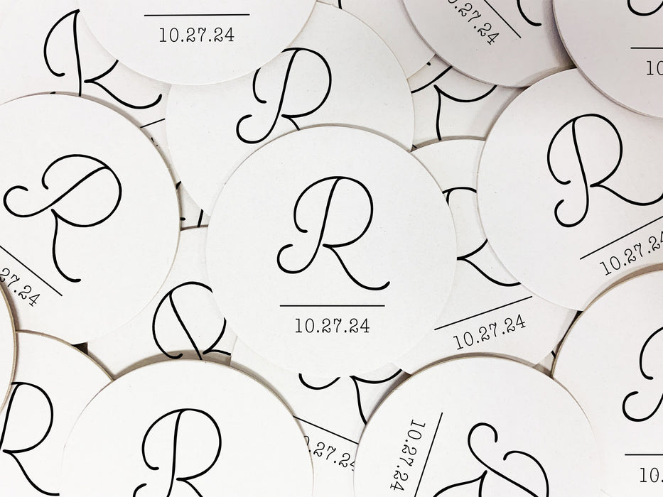 Coasters shown scattered on the surface of a table. Coasters feature Monogrammed Initial design. This design can be customized with a large initial and date in black writing on a plain, white background.