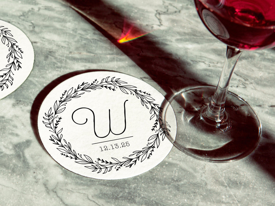 Two coasters are shown with a wine glass on top of one on a gray marble table. Coasters show Monogrammed Floral Wreath Initial design. This design has a floral wreath around a customizable large initial and date, all printed in black.
