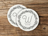 Two coasters are shown stacked on top of each other on wood table. Coasters show Monogrammed Floral Wreath Initial design. This design has a floral wreath around a customizable large initial and date, all printed in black.