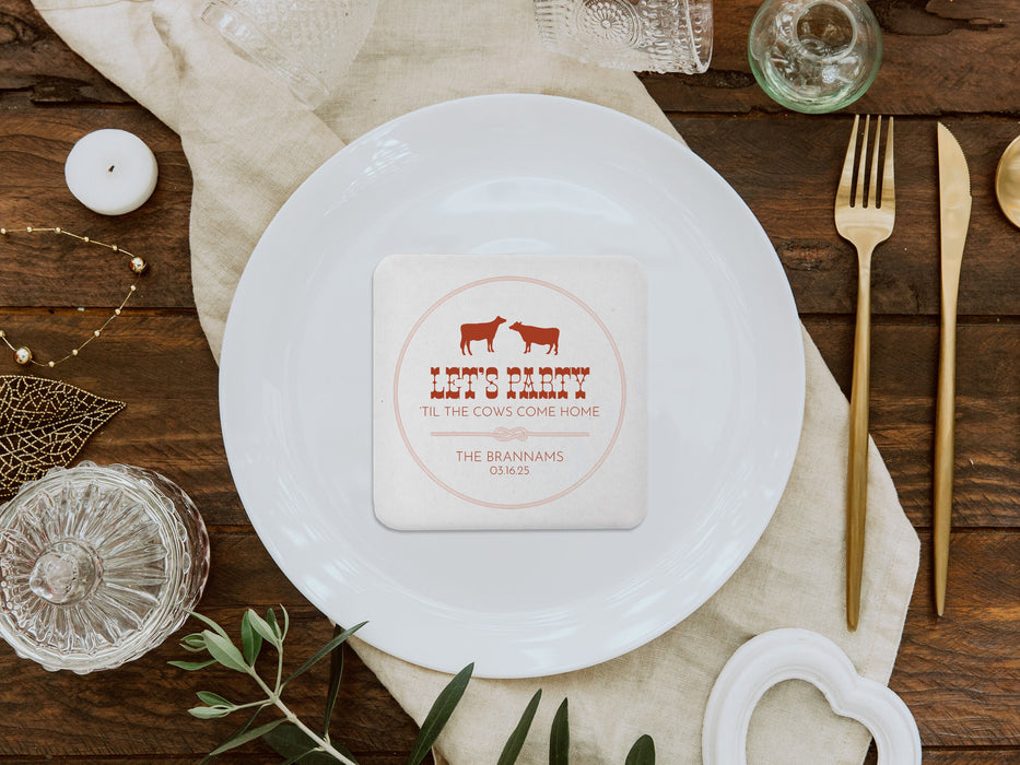 A nice wedding table setting is shown with a coaster in the center of a plate. Coasters feature Til The Cows Come Home design. Design shows two cows with the saying Lets Party Til The Cows Come Home with newlyweds last name and wedding date.