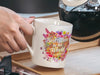 person pouring coffee in a white mug with a floral design with text that says Best Mom in the World est 2023 in a kitchen with utensils and a kettle