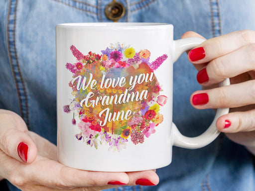 white mug with floral design saying We love you Grandma June being held by woman wearing jean button up shirt and red nail polish