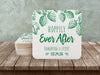 A stack of coasters by a single coaster on a wooden table. Coasters feature Hoppily Ever After design. This design uses green lettering and sketched drawings of beer hops. Wedding couples names and date can be customized.