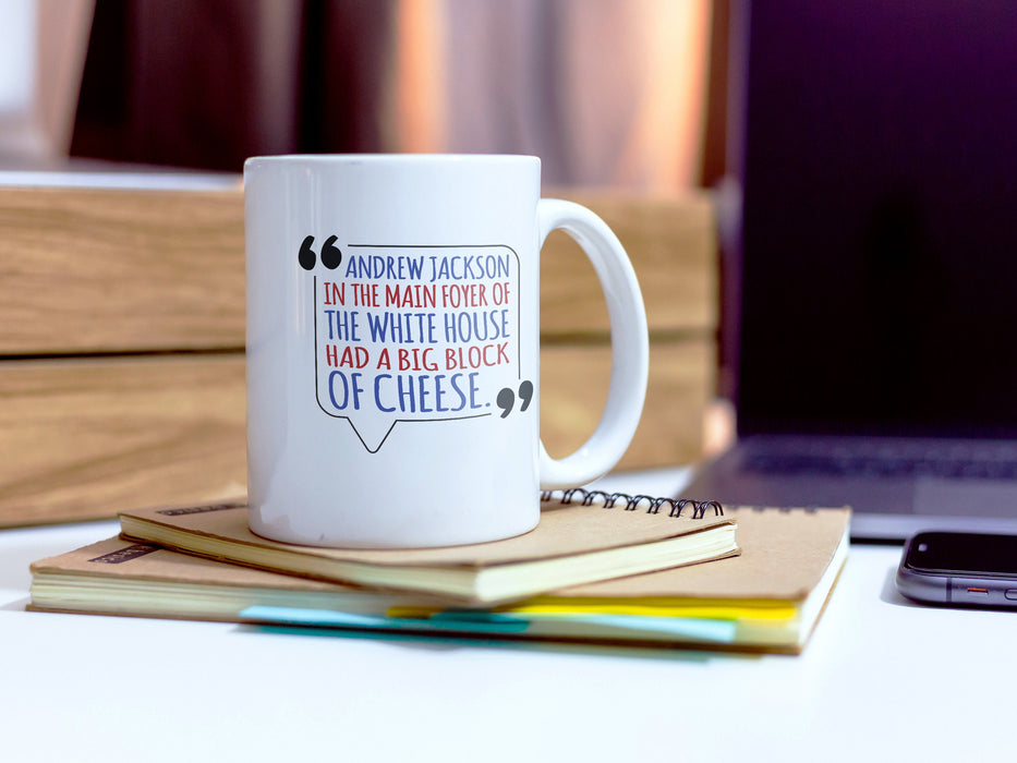 white mug on desk ontop of brown notebooks in front of laptop and phone with American west wing design and Typography that says Andrew Jackson in the Main Foyer of The White House had a Big Block of Cheese.