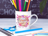 mug with colored pencils in teachers classroom in front of a chalk board surrounded by school materials such as scissors, chalk, notebook, and a blue colored pencil design says Mrs. Johnson 2023