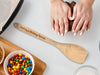 flat wooden spoon on white table next to hands kneading dough and other baking supplies in ceramic and glass bowls as well as a baking pan and cutting board