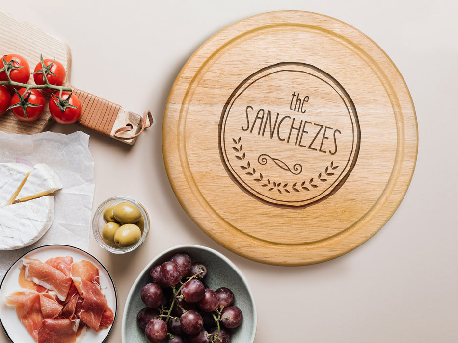 wooden cheeseboard on white table surrounded by charcuterie foods such as Italian meats, olives, grapes, tomatoes and cheese, engraving has The Sanchezes with a circle and wreath design
