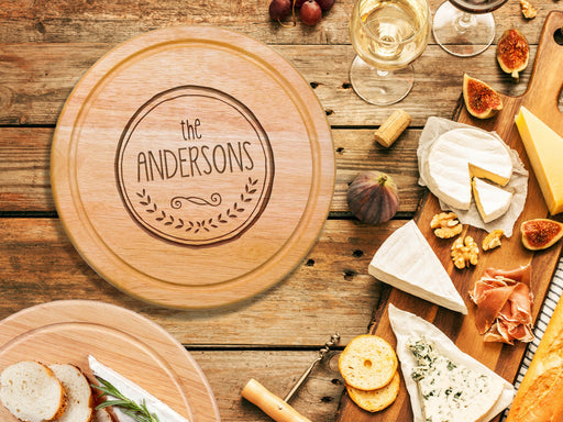 wooden cheeseboard on wooden table surrounded by wine glasses, figs, cheese, bread, crackers, Italian meats, and another charcuterie board engraving includes The Andersons with a circle and wreath design