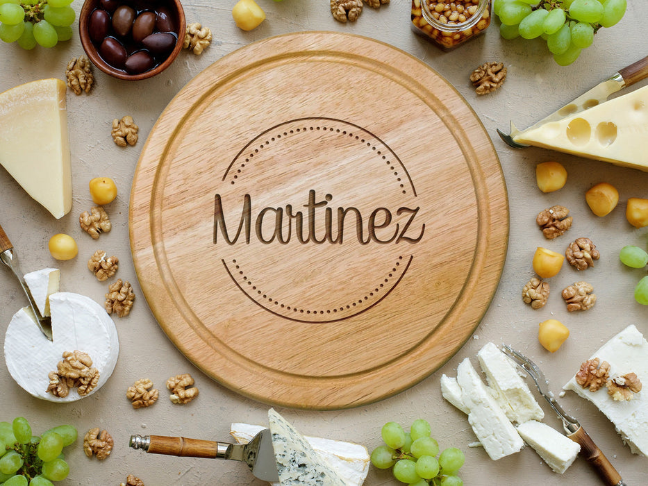 wooden cheese board surrounded by various cheeses, grapes, cheese knife tools, corn nuts, olives and walnuts  engraving is of the name Martinez surrounded by a circle and dots design