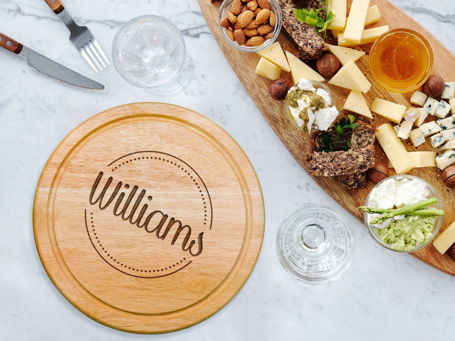 cheeseboard on marble table surrounded by tableware such as glasses, forks, knives, cheese, nuts, dips, and other charcuterie foods engraving includes the name Williams surrounded by dots and circle design