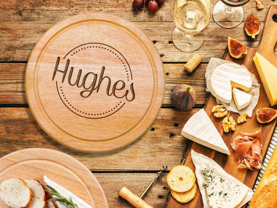wooden cheeseboard on wooden table surrounded by wine glasses, figs, cheese, bread, crackers, Italian meats, and another charcuterie board engraving includes the name Hughs surrounded by a circle and dots design