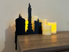 A black door frame topper, designed as a silhouette of three dripping wax candles, is seen on a wooden table surface with candles sat behind and in front of it. The wall behind is a grey blue.