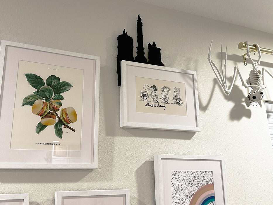 A photo collage wall is shown with various artworks in each frame. A black door frame topper, designed as a silhouette of three dripping wax candles, is on top of one of the white picture frames. The wall behind is also white.