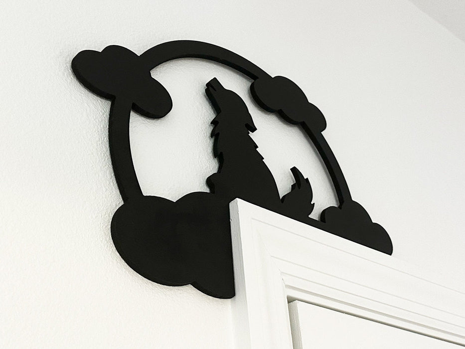 A black door frame topper, designed with the silhouette of a howling wolf, full moon, and clouds, is seen on top of a white door frame. The wall behind is white.