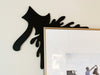 A black door frame topper, designed with the silhouette of an axe and blood splashes, is seen on top of a picture frame. The wall behind is beige.