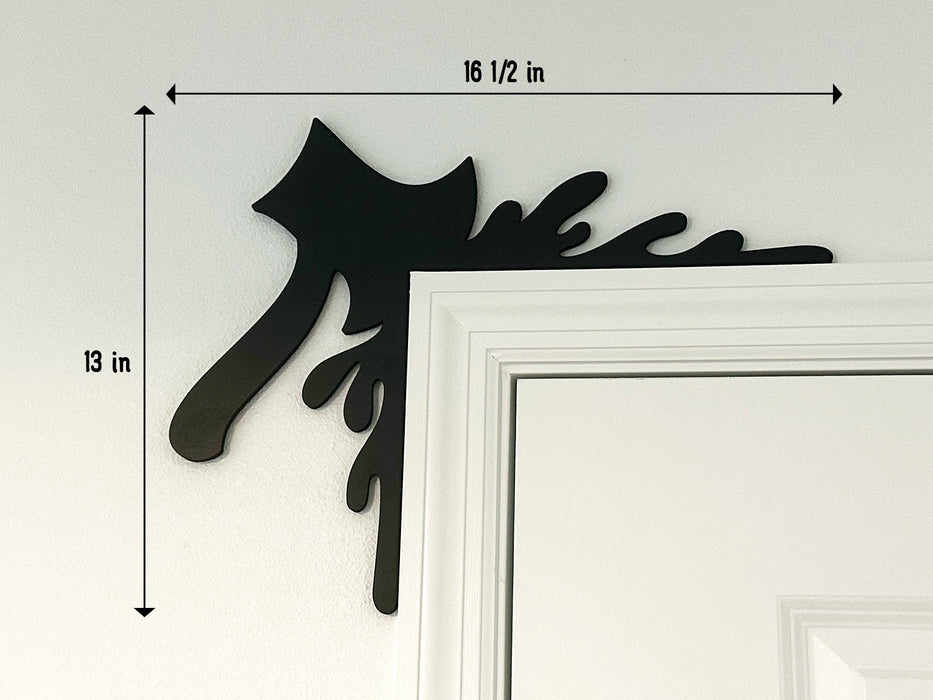 A black door frame topper, designed with the silhouette of an axe and blood splashes, is seen on top of a white door frame. The topper measures about 16 1/2 inches wide by 13 inches tall. The wall behind is white.