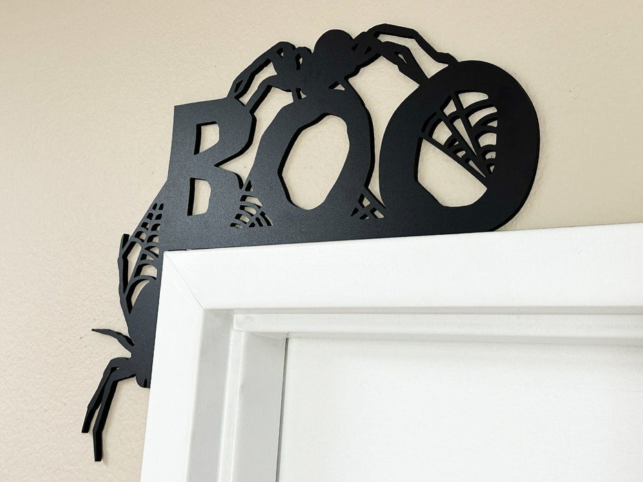 A black door frame topper, designed with the word BOO, spiders, and spider webs, is seen on top of a white door frame. The wall behind is beige.
