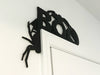 A black door frame topper, designed with the word BOO, spiders, and spider webs, is seen on top of a white door frame. The wall behind is white.