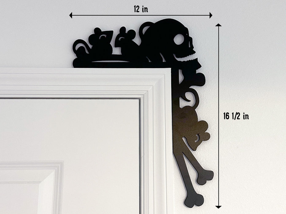 A black door frame topper, designed with the silhouette of scurrying rats, bones, and a skull, is seen on top of a white door frame. The topper measures about 12 inches wide by 16 1/2 inches tall. The wall behind is white.