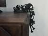 A black door frame topper, designed with the silhouette of scurrying rats, bones, and a skull, is seen on top of a wooden entertainment center. The wall behind is grey.