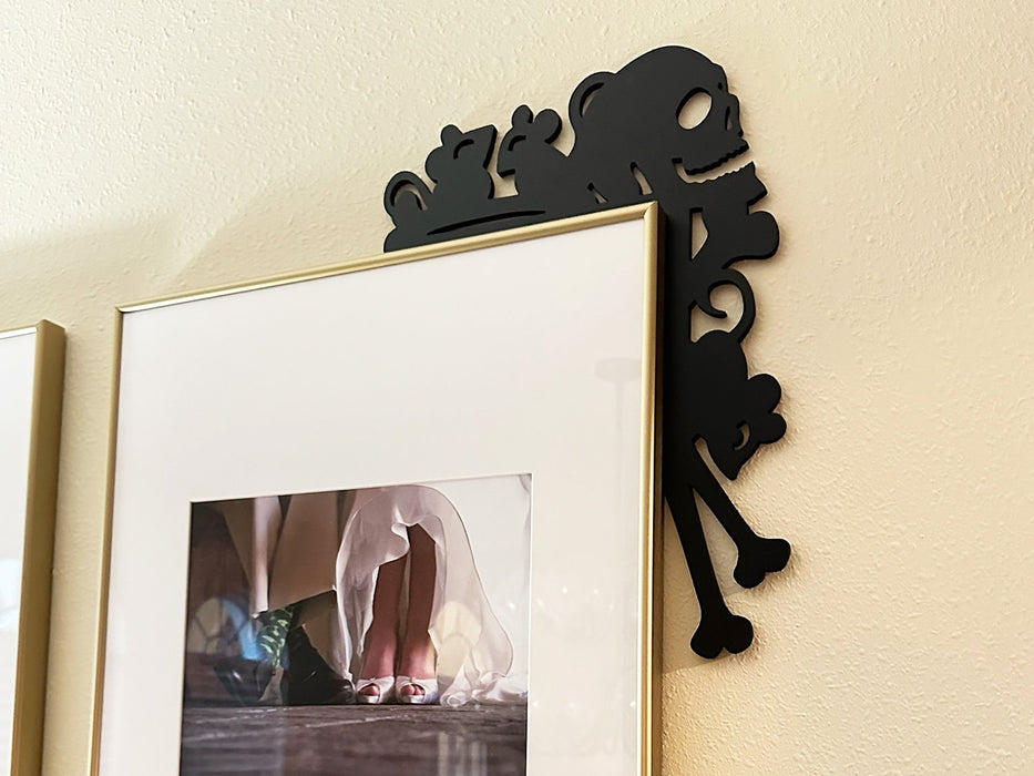 A black door frame topper, designed with the silhouette of scurrying rats, bones, and a skull, is seen on top of a picture frame. The wall behind is beige.