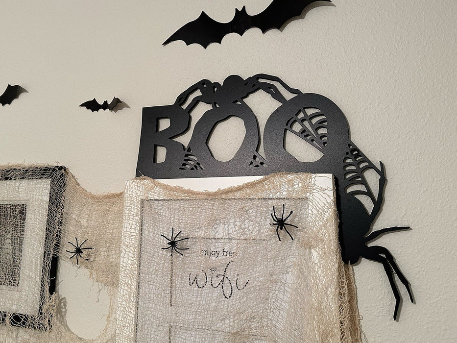 A black door frame topper, designed with the word BOO, spiders, and spider webs, is seen on top of white picture frame. Spooky spider webs and spiders hang across the frame. A beige wall seen behind the frame has fake bats attached to it.