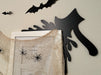 A black door frame topper, designed with the silhouette of an axe and blood splashes, is seen on top of a white picture frame. The frame is covered in spider webs and fake spiders. The beige wall behind has fake bats attached to it.