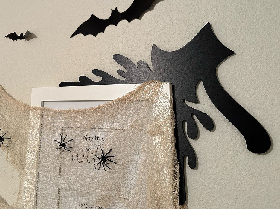 A black door frame topper, designed with the silhouette of an axe and blood splashes, is seen on top of a white picture frame. The frame is covered in spider webs and fake spiders. The beige wall behind has fake bats attached to it.