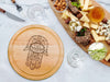 cheeseboard on marble table surrounded by tableware such as glasses, forks, knives, cheese, nuts, dips, and other charcuterie foods engraving includes a hamsa design