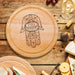 wooden cheeseboard on wooden table surrounded by wine glasses, figs, cheese, bread, crackers, Italian meats, and another charcuterie board engraving includes a Hamsa design