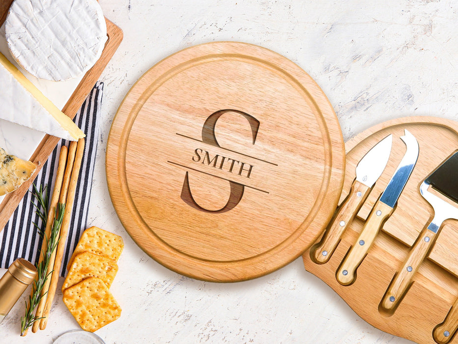 wooden cheeseboard on marble table open with stainless steel cheese knives tool set surrounded by cheeses, breads, and crackers engraving has the letter S with the name Smith on it