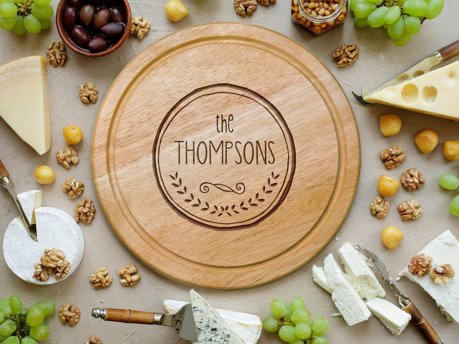 wooden cheese board surrounded by various cheeses, grapes, cheese knife tools, corn nuts, olives and walnuts  engraving is of The Thompsons with a circle and wreath design