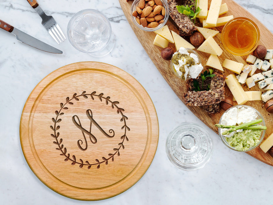 cheeseboard on marble table surrounded by tableware such as glasses, forks, knives, cheese, nuts, dips, and other charcuterie foods engraving includes the initial M surrounded by a wreath design
