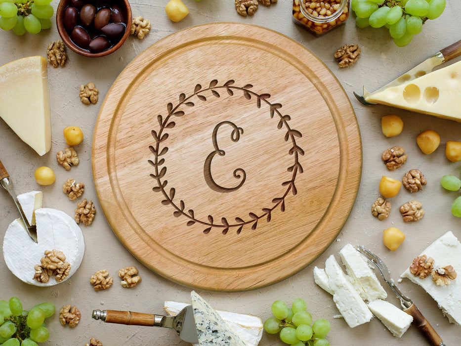 wooden cheese board surrounded by various cheeses, grapes, cheese knife tools, corn nuts, olives and walnuts  engraving is of the initial E surrounded by a wreath design