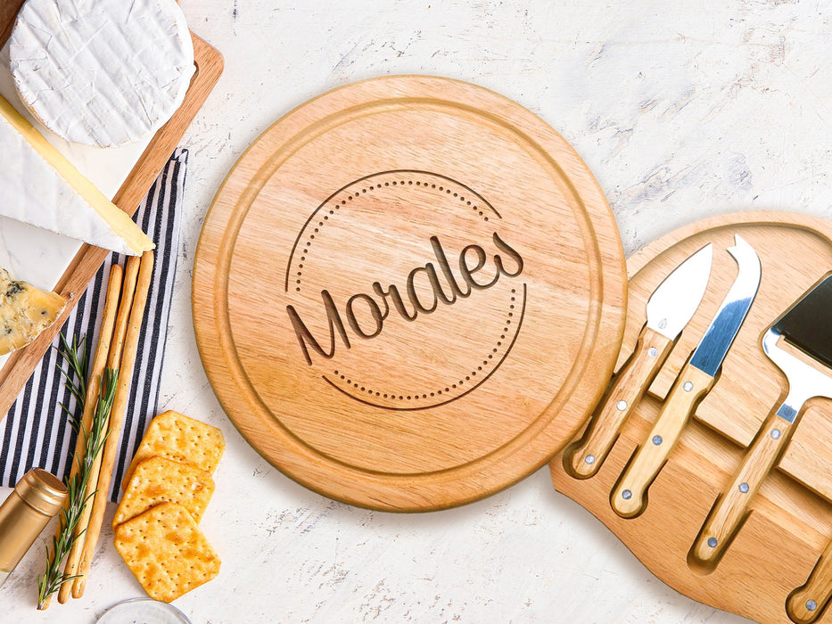 wooden cheeseboard on marble table open with stainless steel cheese knives tool set surrounded by cheeses, breads, and crackers engraving has the name Morales surrounded by a circle and dots design