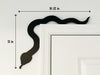 A black door frame topper, designed with the silhouette of a slithering snake, is seen on top of a white door frame. The topper measures about 16 1/2 inches wide by 13 inches tall. The wall behind is white.