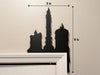 A black door frame topper, designed as a silhouette of three dripping wax candles, is seen on top of a white door frame. The topper measures about 9 inches wide by 14 inches tall. The wall behind is beige.