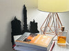 A black door frame topper, designed as a silhouette of three dripping wax candles, is seen on a wooden table surface with a lamp, a stack of books, and perfume besides it. The wall behind is a grey.