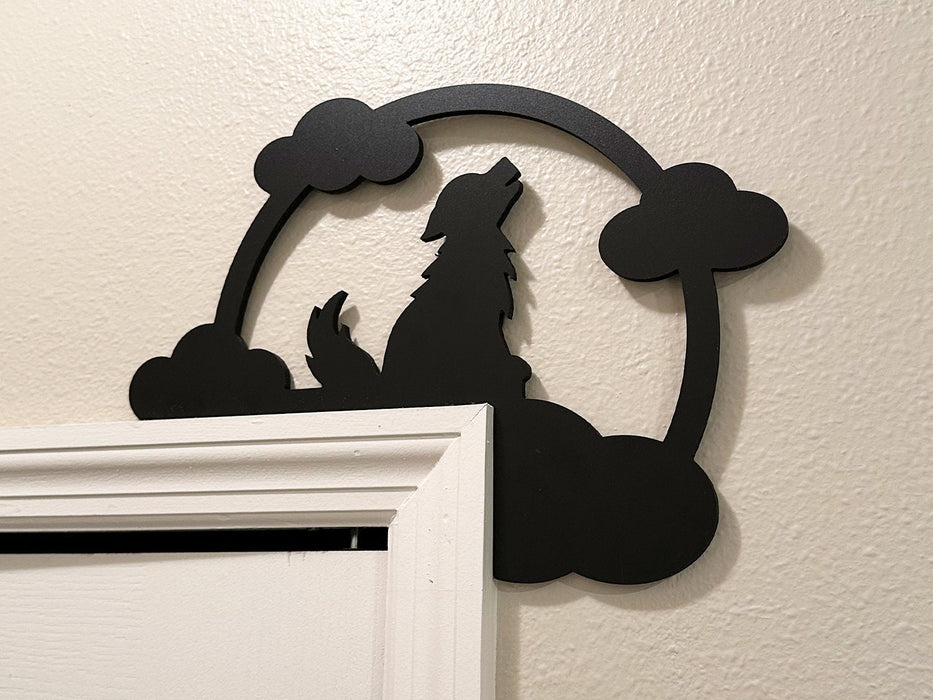 A black door frame topper, designed with the silhouette of a howling wolf, full moon, and clouds, is seen on top of a white door frame. The wall behind is beige.