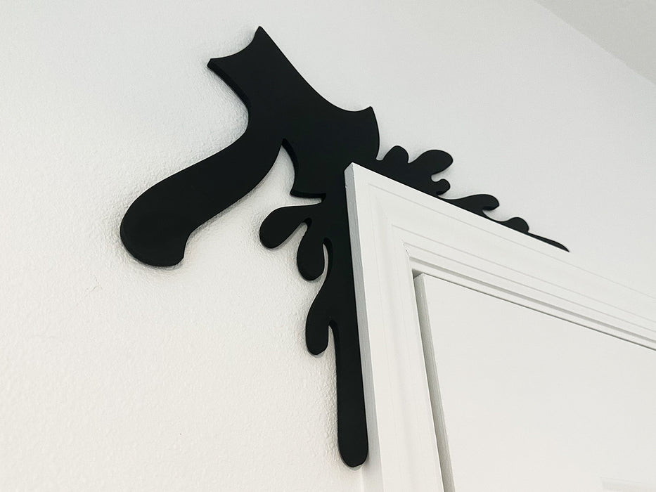 A black door frame topper, designed with the silhouette of an axe and blood splashes, is seen on top of a white door frame. The wall behind is white.
