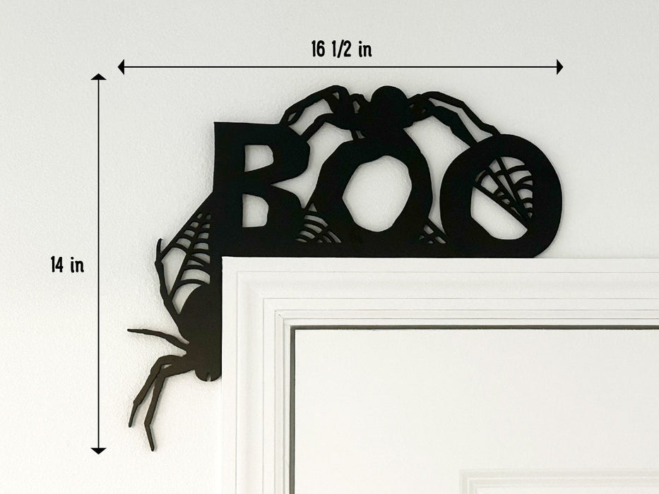 A black door frame topper, designed with the word BOO, spiders, and spider webs, is seen on top of a white door frame. The topper measures about 16 1/2 inches wide by 14 inches tall. The wall behind is white.