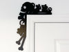 A black door frame topper, designed with the silhouette of scurrying rats, bones, and a skull, is seen on top of a white door frame. The wall behind is white.