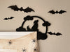 A black door frame topper, designed with the silhouette of a howling wolf, full moon, and clouds, is seen on top of a white picture frame. The frame is covered in spider webs and fake spiders. The beige wall behind has fake bats attached to it.