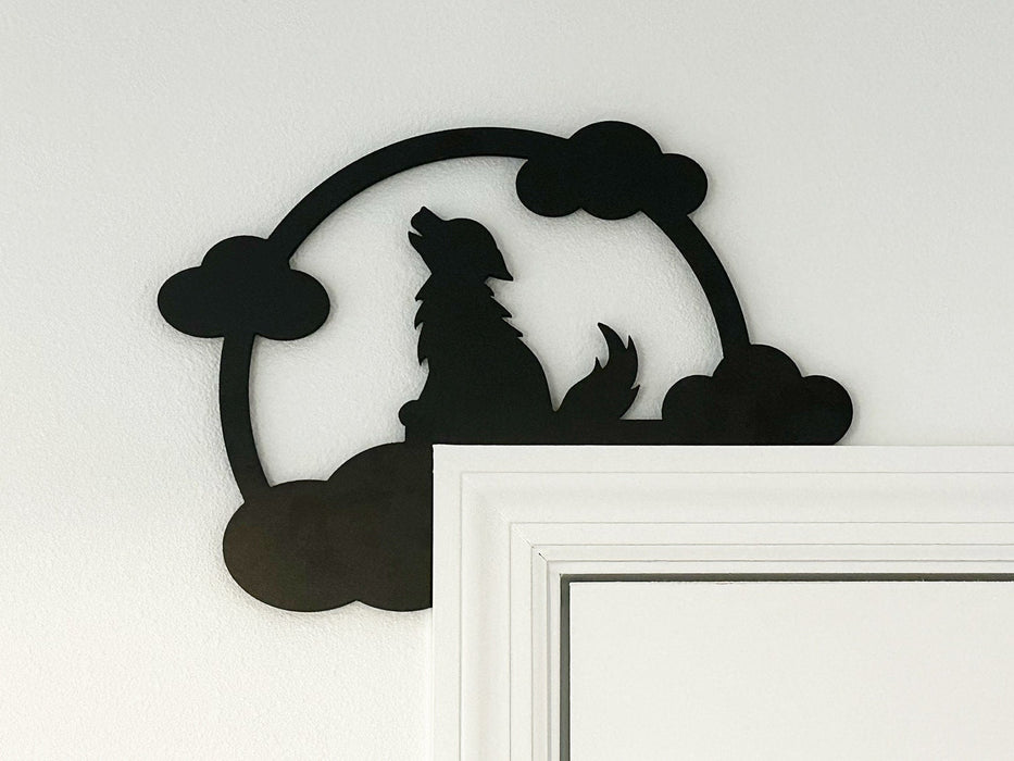 A black door frame topper, designed with the silhouette of a howling wolf, full moon, and clouds, is seen on top of a white door frame. The wall behind is white.