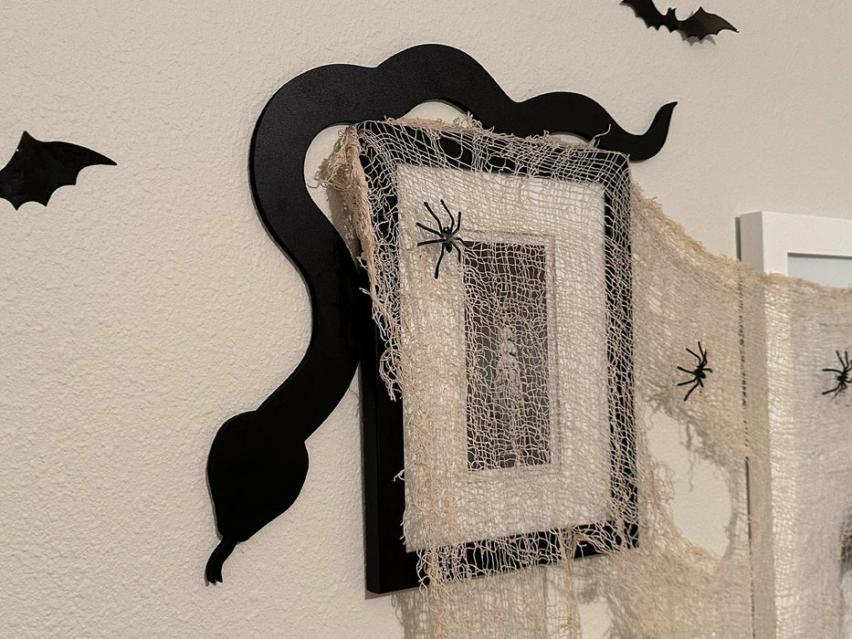 A black door frame topper, designed with the silhouette of a slithering snake, is seen on top of a black picture frame that's covered in spider webs and fake spiders. The beige wall behind is decorated with fake bats.