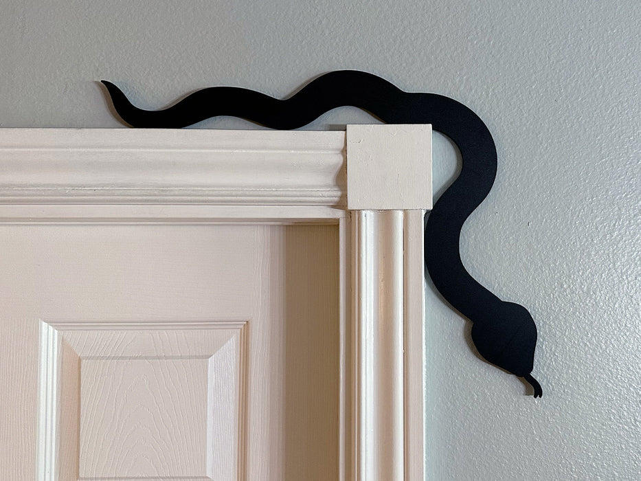 A black door frame topper, designed with the silhouette of a slithering snake, is seen on top of a white door frame. The wall behind is blue.