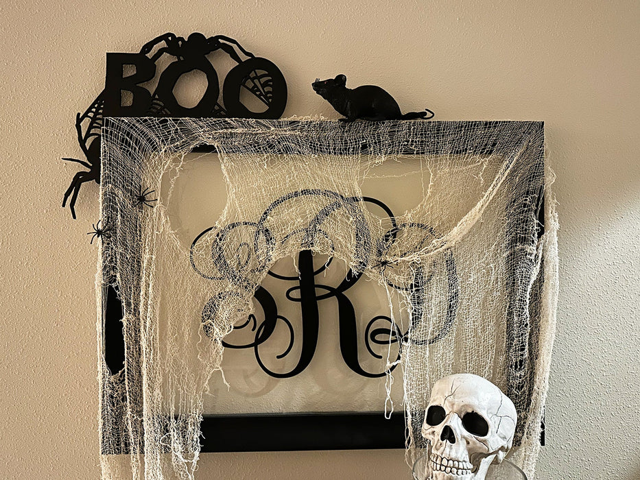 A black door frame topper, designed with the word BOO, spiders, and spider webs, is seen on top of black frame. The frame is covered in spider webs. A fake rat, skull, and spiders can be seen on the frame. The wall behind is beige.
